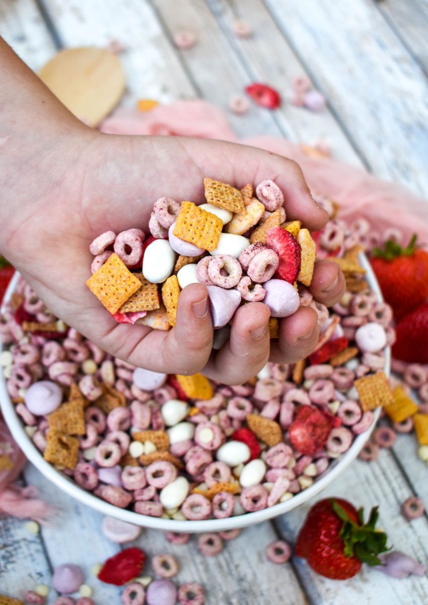 a hand holding a scoop of strawberry snack mix above a white mixing bowl