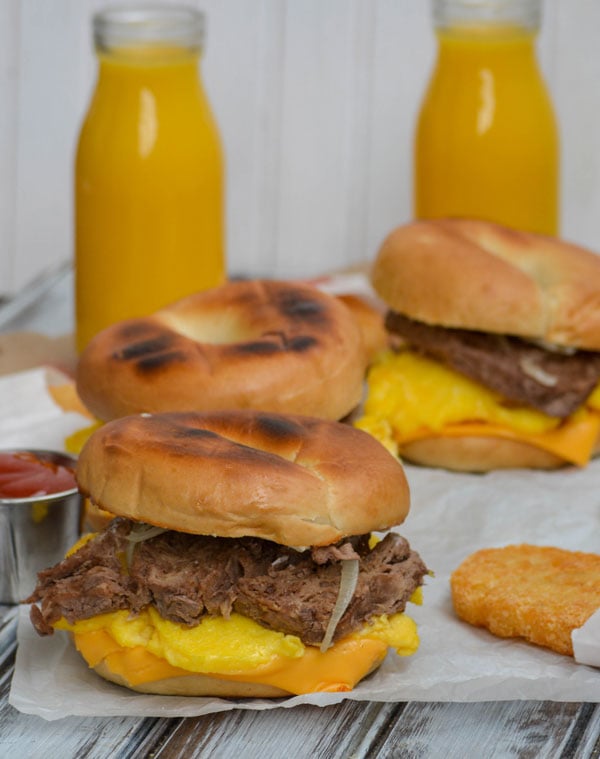 copycat steak egg and cheese bagel breakfast sandwiches on a sheet of crinkled white parchment paper with glasses of orange juice
