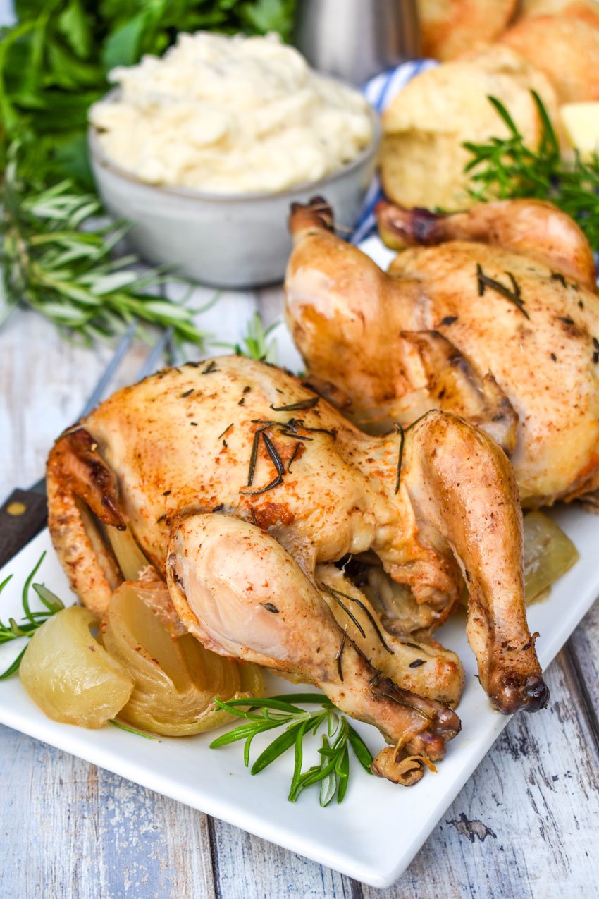 two crockpot cornish game hens on a white serving platter surrounded by cooked onions & fresh herbs