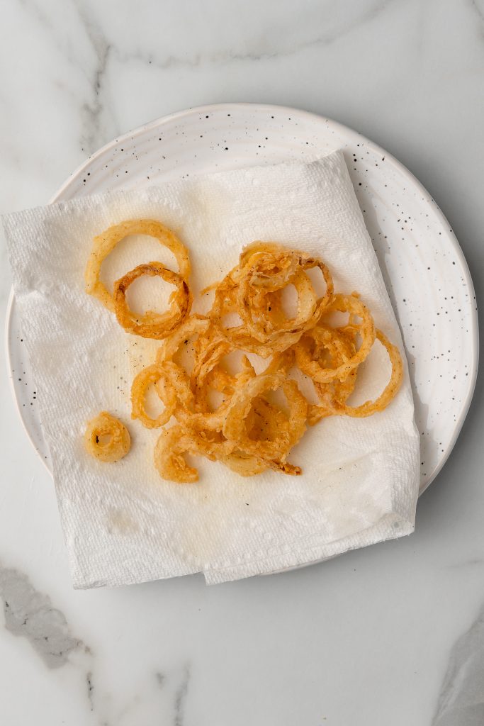 freshly fried crispy onions straws resting on a paper towel lined white plate