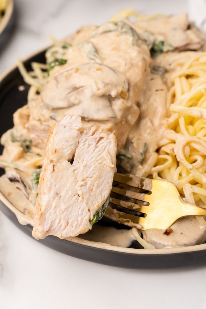 a gold fork next to a piece of creamy champagne chicken on top of pasta on a black plate