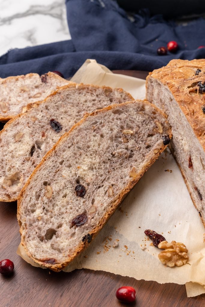 a slice of no knead cranberry walnut bread on a parchment paper lined wooden cutting board