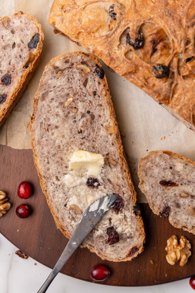 a silver knife spreading softened butter on a slice of no knead cranberry walnut bread