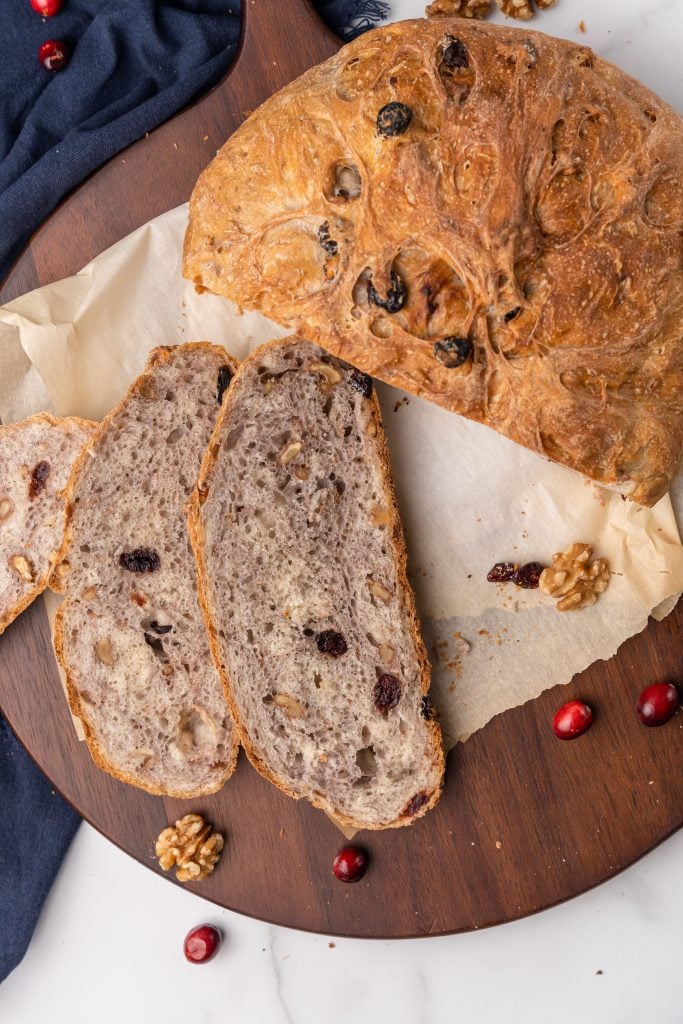 a sliced loaf of cranberry walnut dutch oven bread on a parchment paper lined wooden cutting board