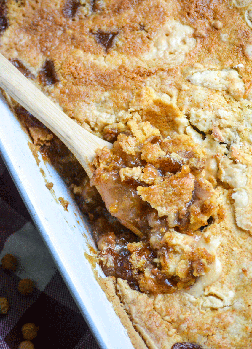a wooden spoon digging into a caramel apple pie cake in a white casserole dish