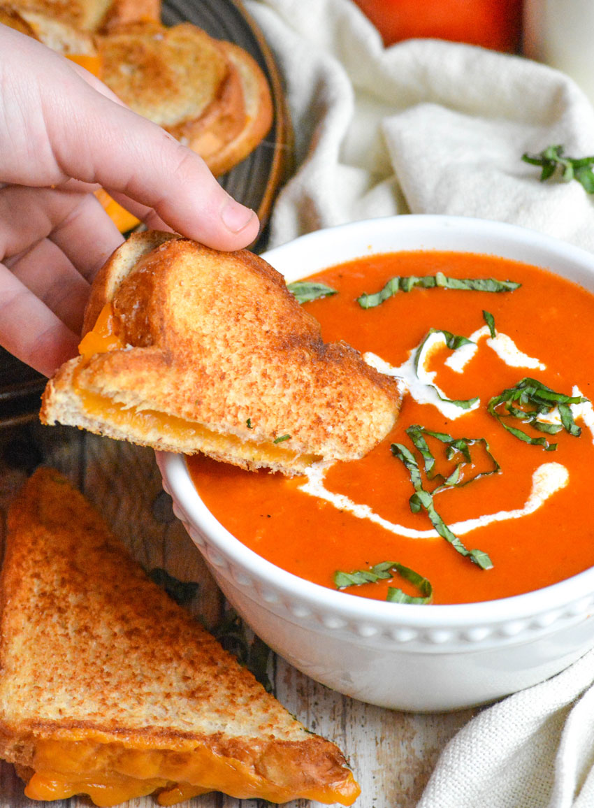 a hand dipping a grilled cheese sandwich into a bowl of panera bread tomato basil soup