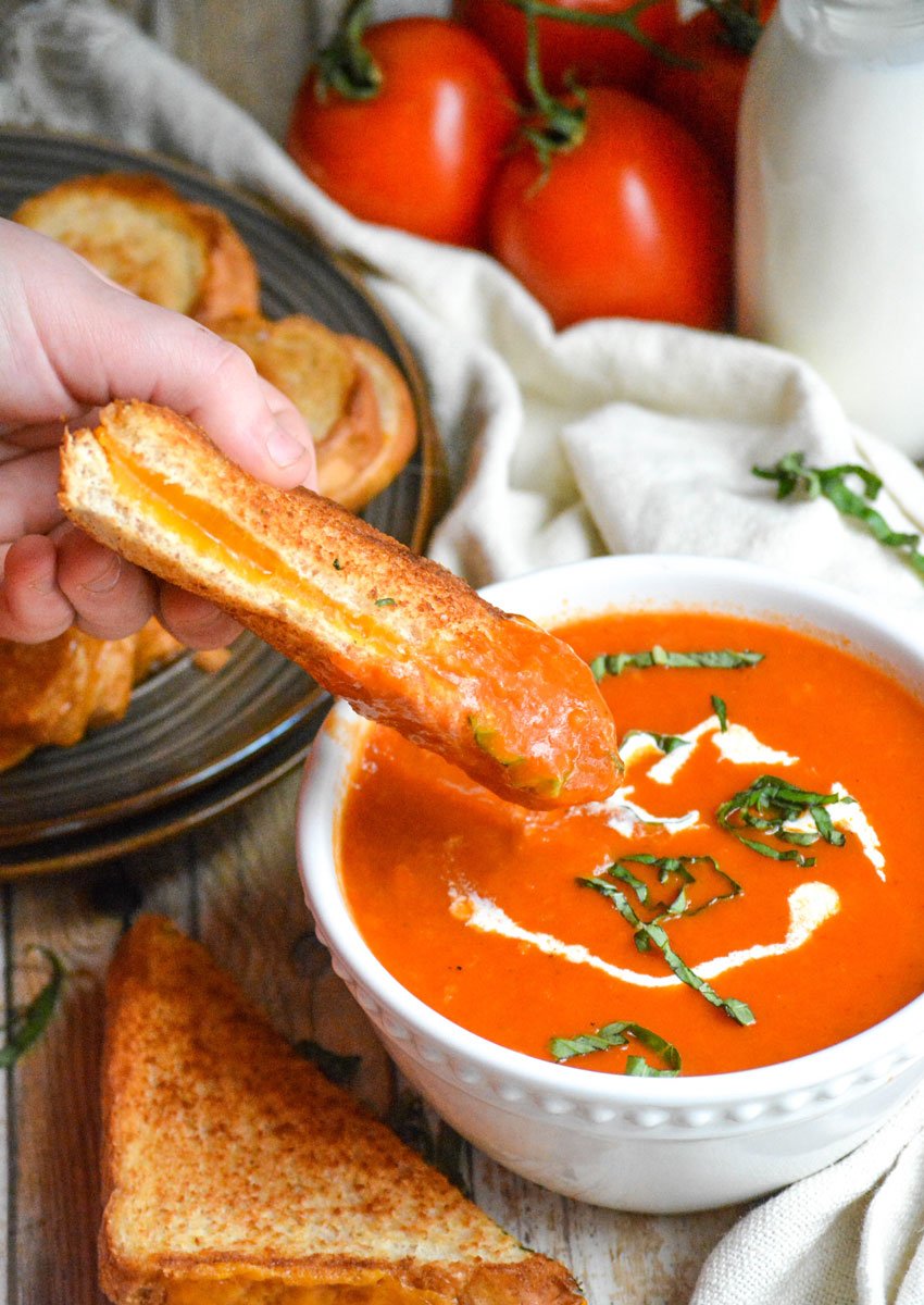 a hand dipping a grilled cheese sandwich into a bowl of panera bread tomato basil soup