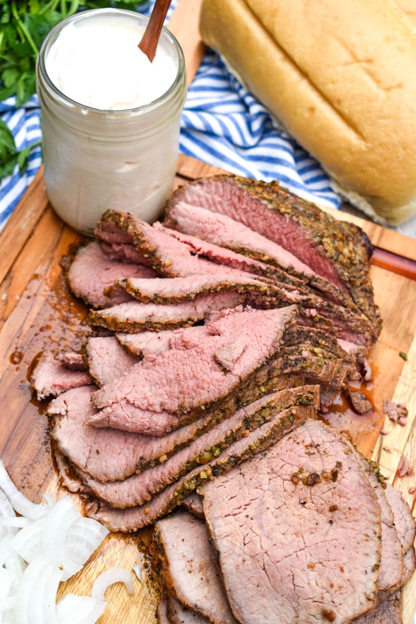 slices of medium smoked eye of round roast on a white cutting board surrounded by the ingredients needed to make smoked roast beef sandwiches