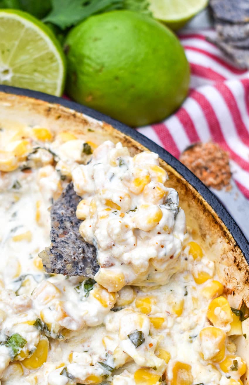 a tortilla chip resting on the edge of a skillet topped with scoop of cheesy hot corn dip