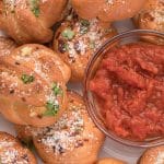 homemade garlic knots piled on a white serving plate around a small glass bowl filled with tomato dipping sauce