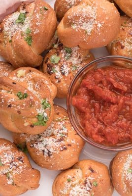 homemade garlic knots piled on a white serving plate around a small glass bowl filled with tomato dipping sauce