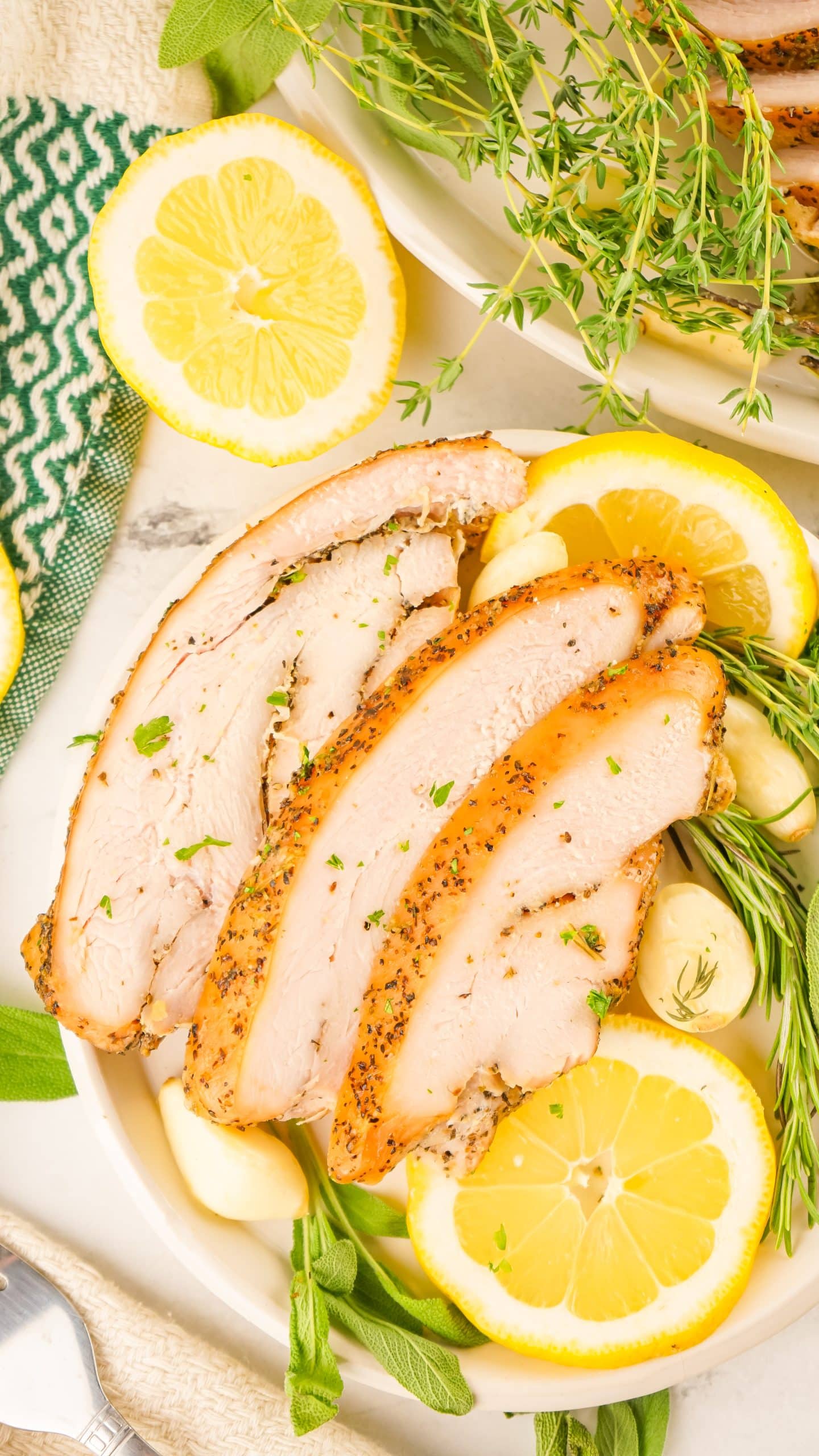 slices of a smoked turkey breast on a bed of fresh herbs and sliced lemons on a white platter
