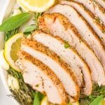 slices of a smoked turkey breast on a bed of fresh herbs and sliced lemons on a white platter