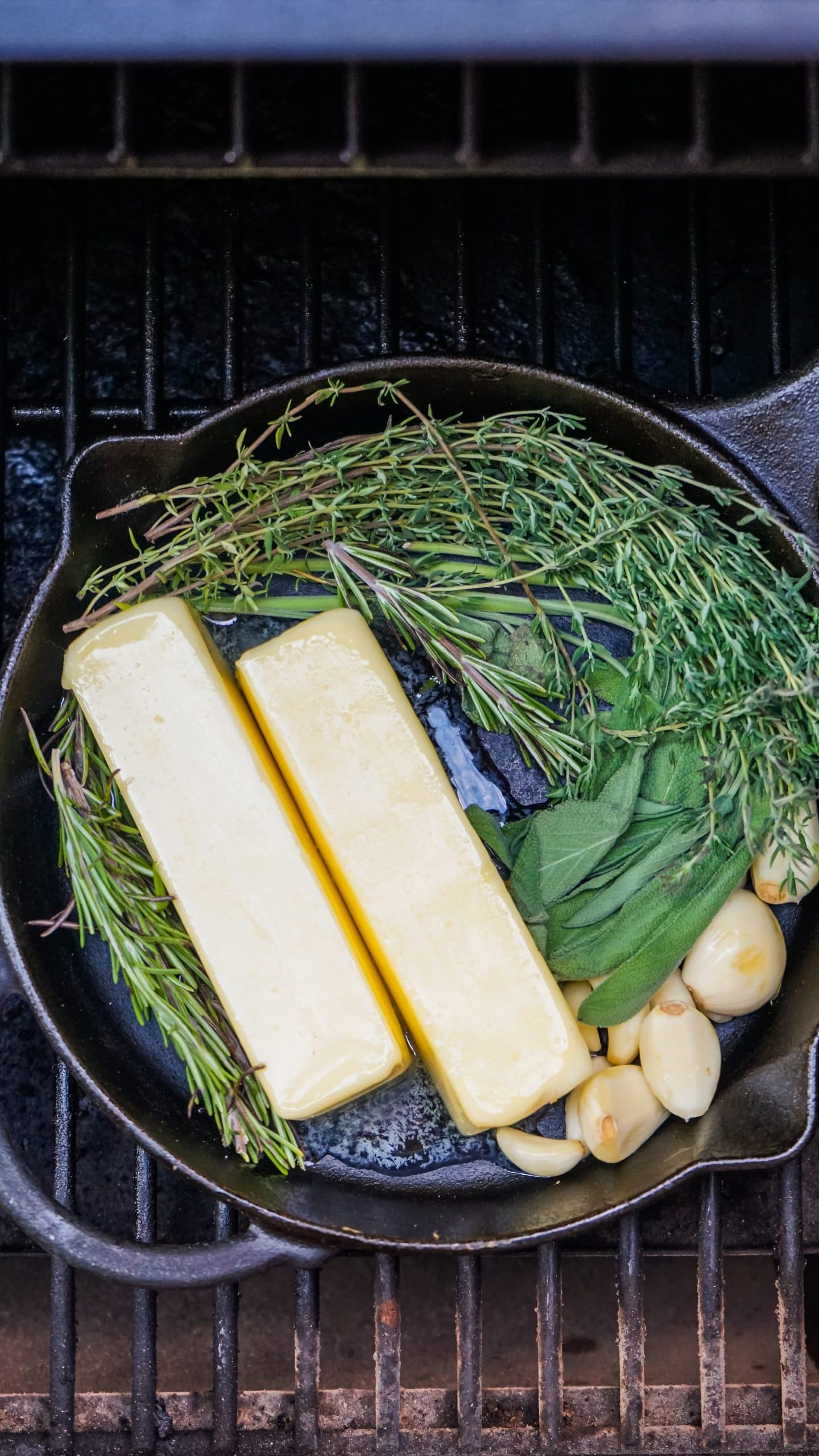 sticks of butter and fresh herbs in a cast iron skillet sitting on smoker grates