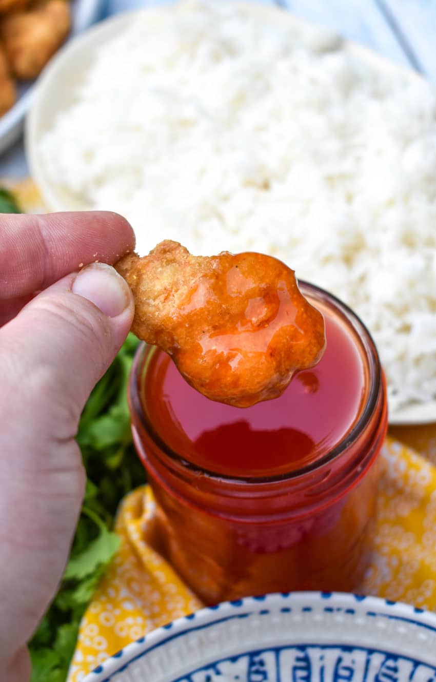 a hand holding up a piece of sweet and sour chicken dipped in red sauce