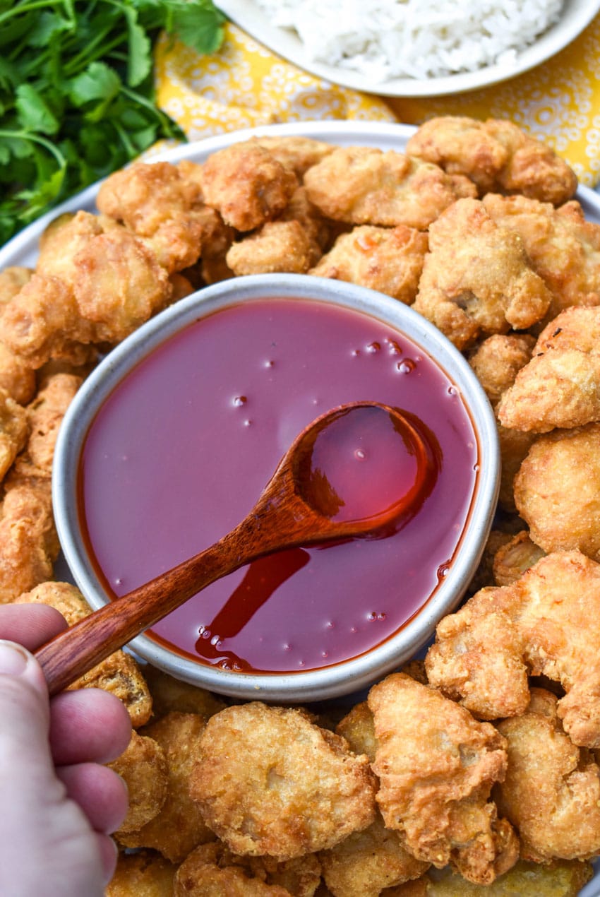 a wooden spoon in a gray bowl filled with sweet and sour sauce surrounded by crispy pieces of chicken