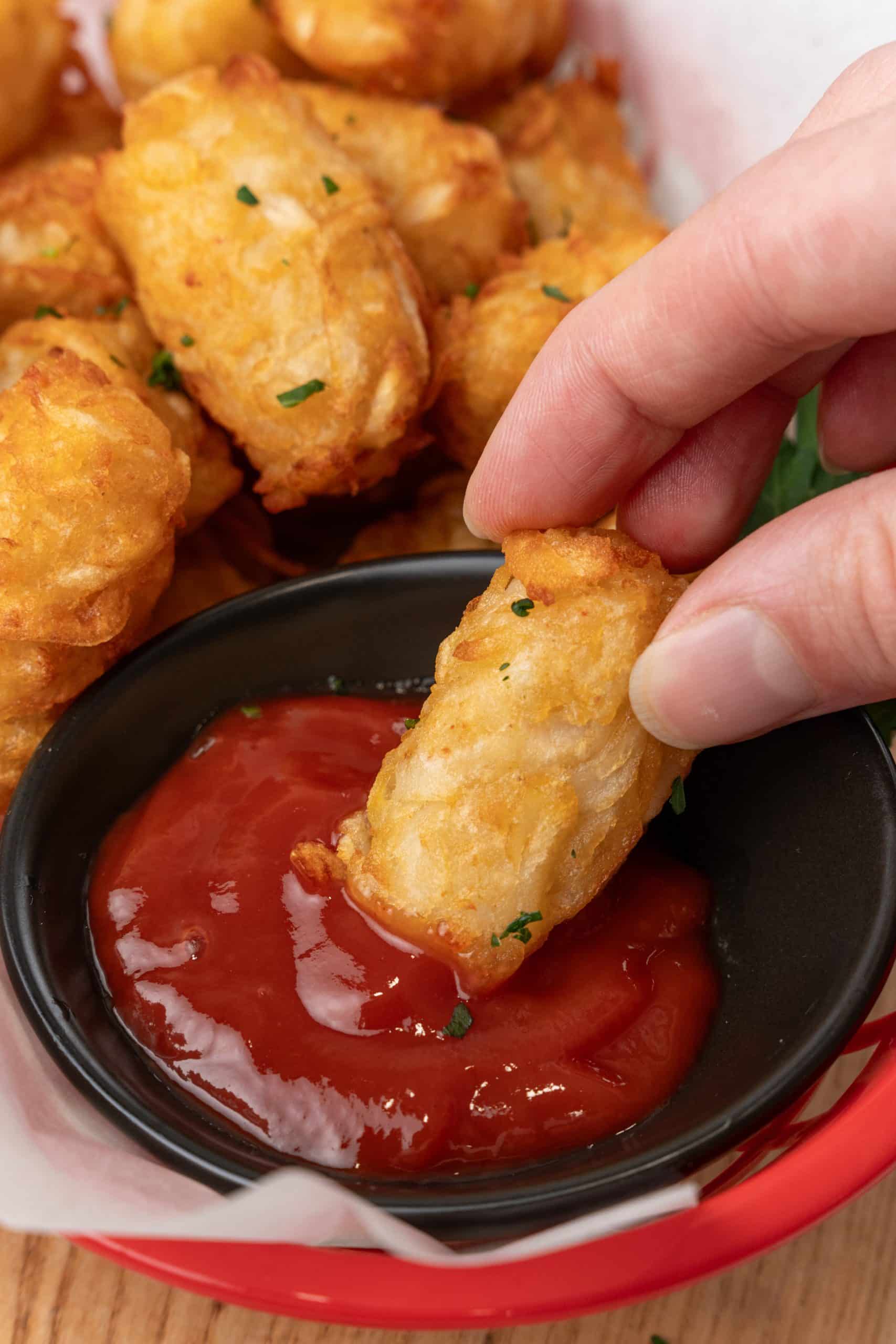 a hand dunking a fried tater tot in a small black bowl filled with ketchup