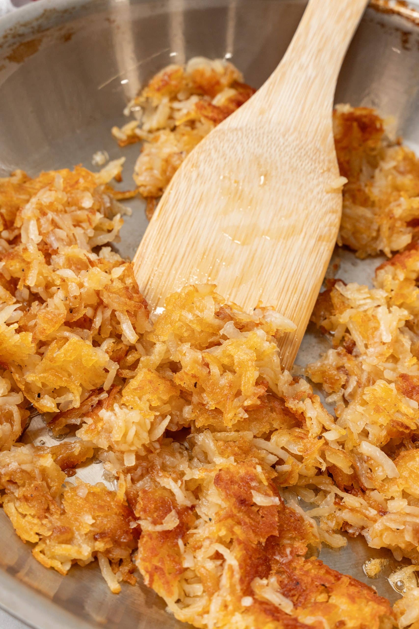 a wooden spatula stirring crispy golden hash browns in a silver skillet