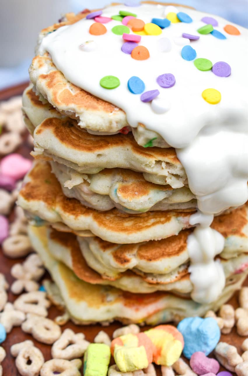 a stack of lucky charms pancakes with white chocolate drizzled overtop on a wooden cutting board with cereal on the sides