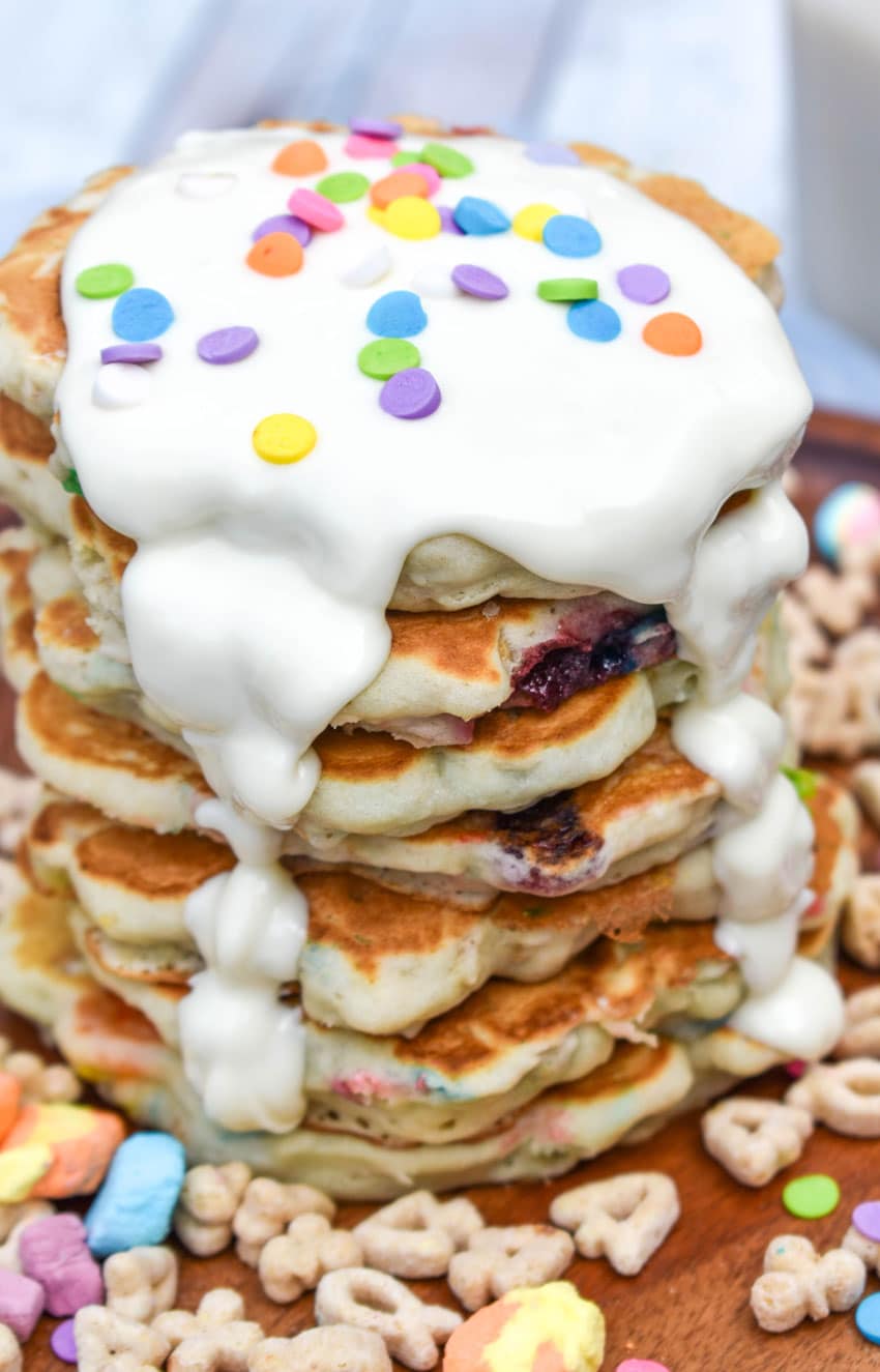 a stack of lucky charms pancakes with white chocolate drizzled overtop on a wooden cutting board with cereal on the sides
