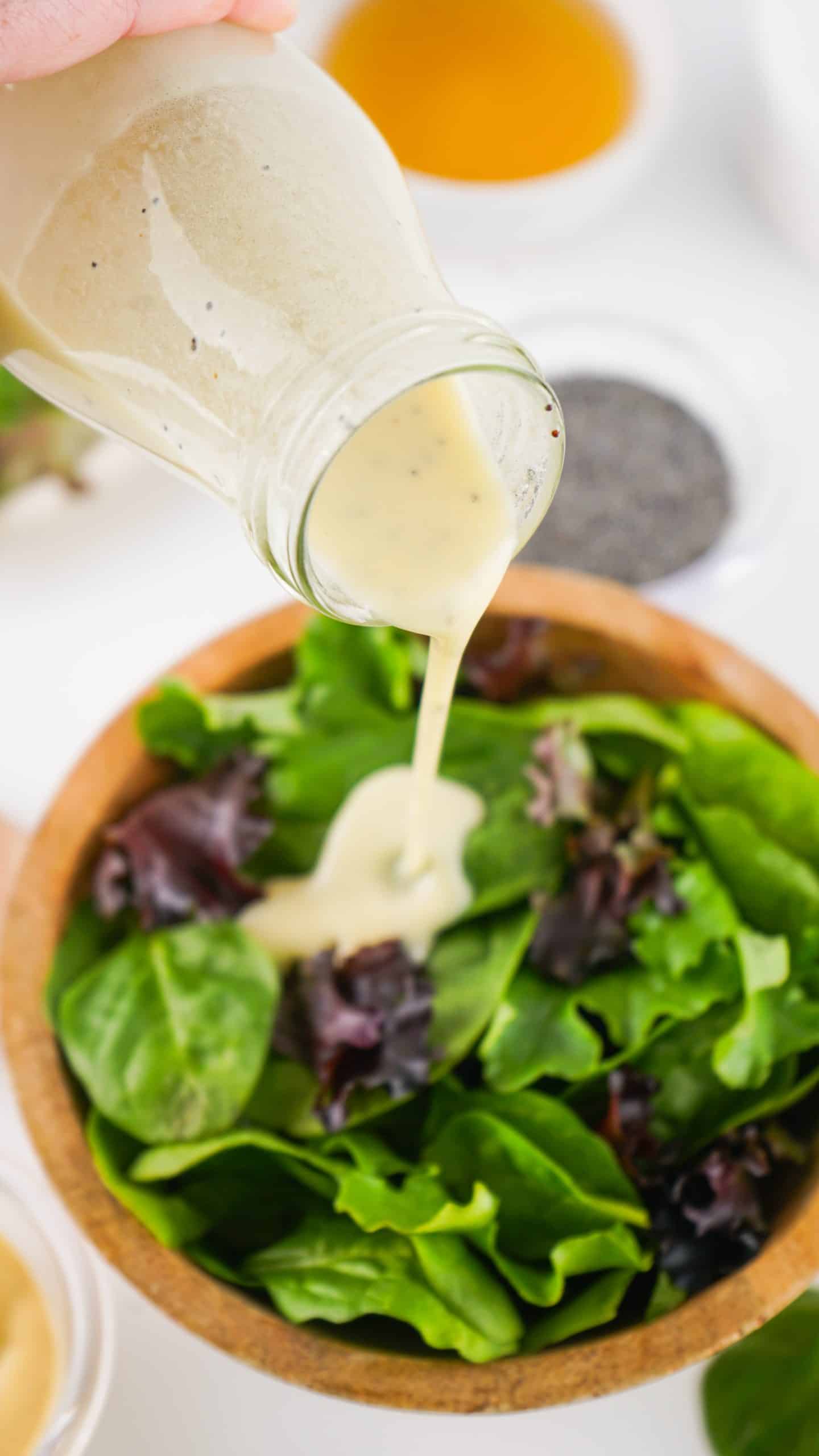 poppy seed dressing recipe being poured out of a glass jar over salad greens in a wooden bowl