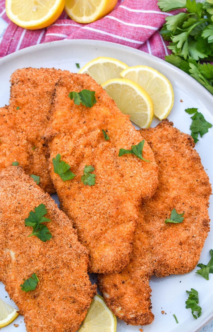 air fryer chicken cutlets in a pile on a white plate with fresh herbs sprinkled on top and lemon slices on the side