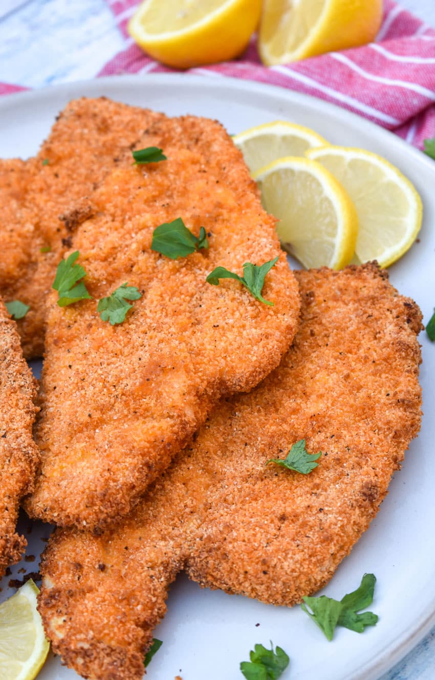 air fryer chicken cutlets in a pile on a white plate with fresh herbs sprinkled on top and lemon slices on the side