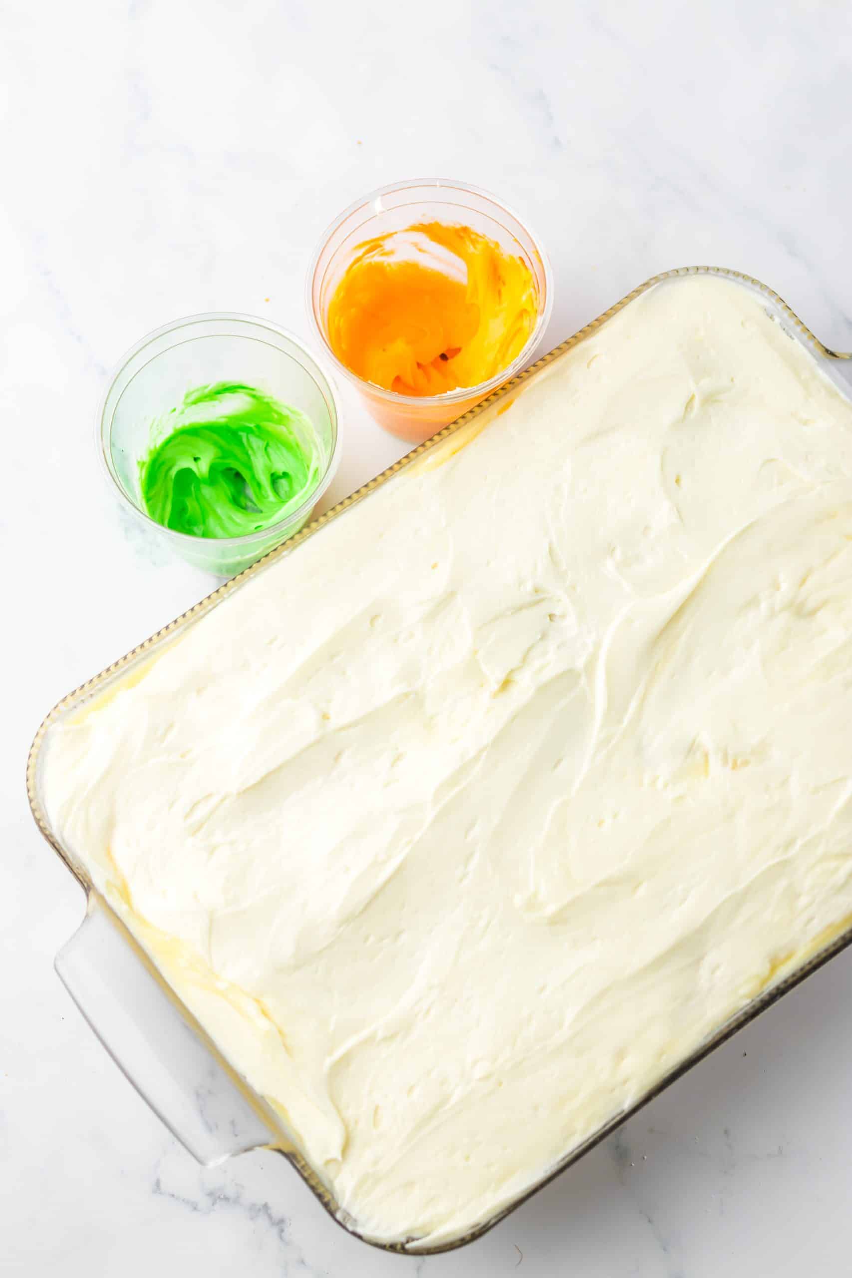 a vanilla frosted carrot cake with cups of orange and green dyed frosting on the side