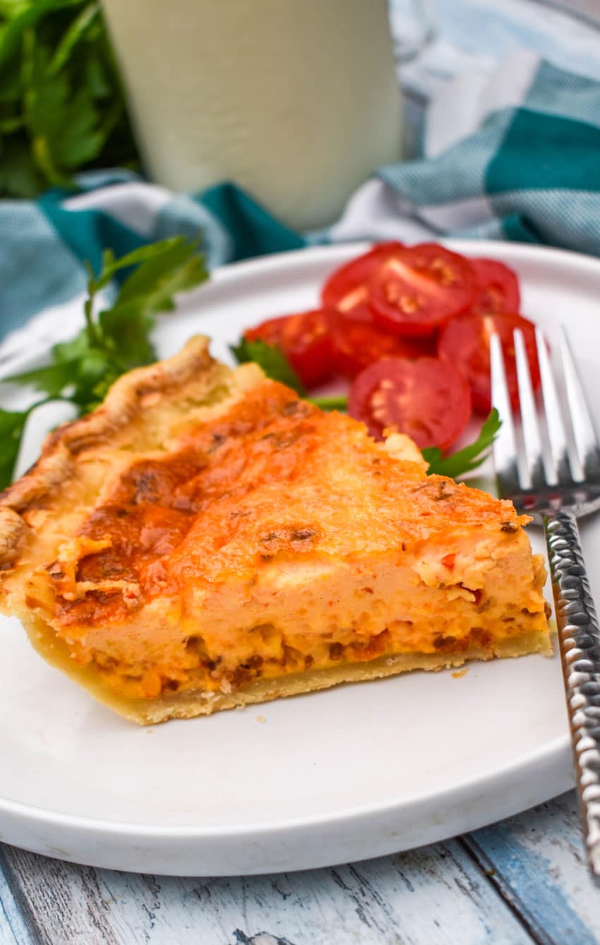 a slice of pimento cheese and bacon quiche on a white plate with sliced tomatoes and a silver fork on the side
