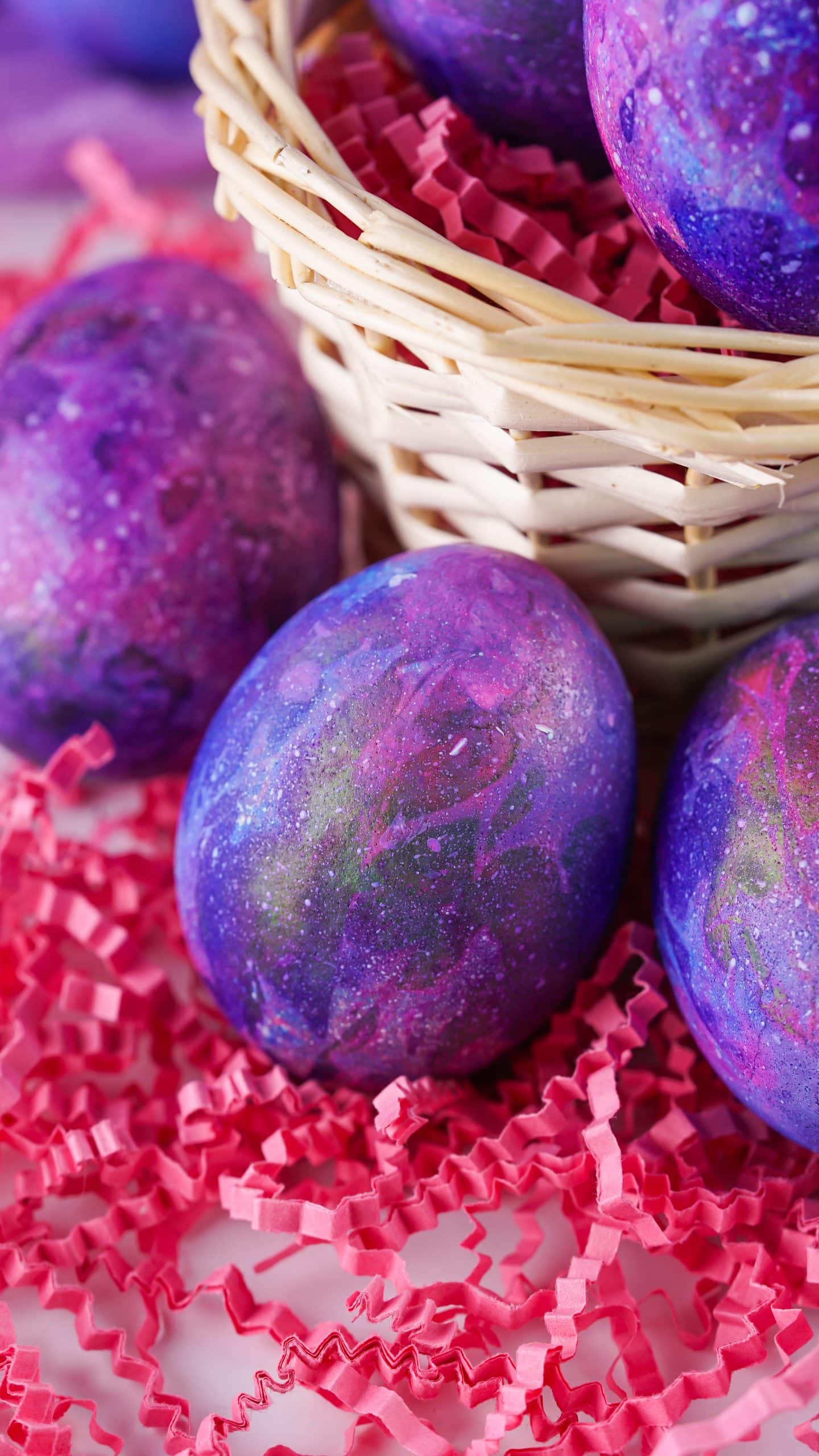 space colored dyed easter eggs resting on red crumpled strings of paper next to a straw basket