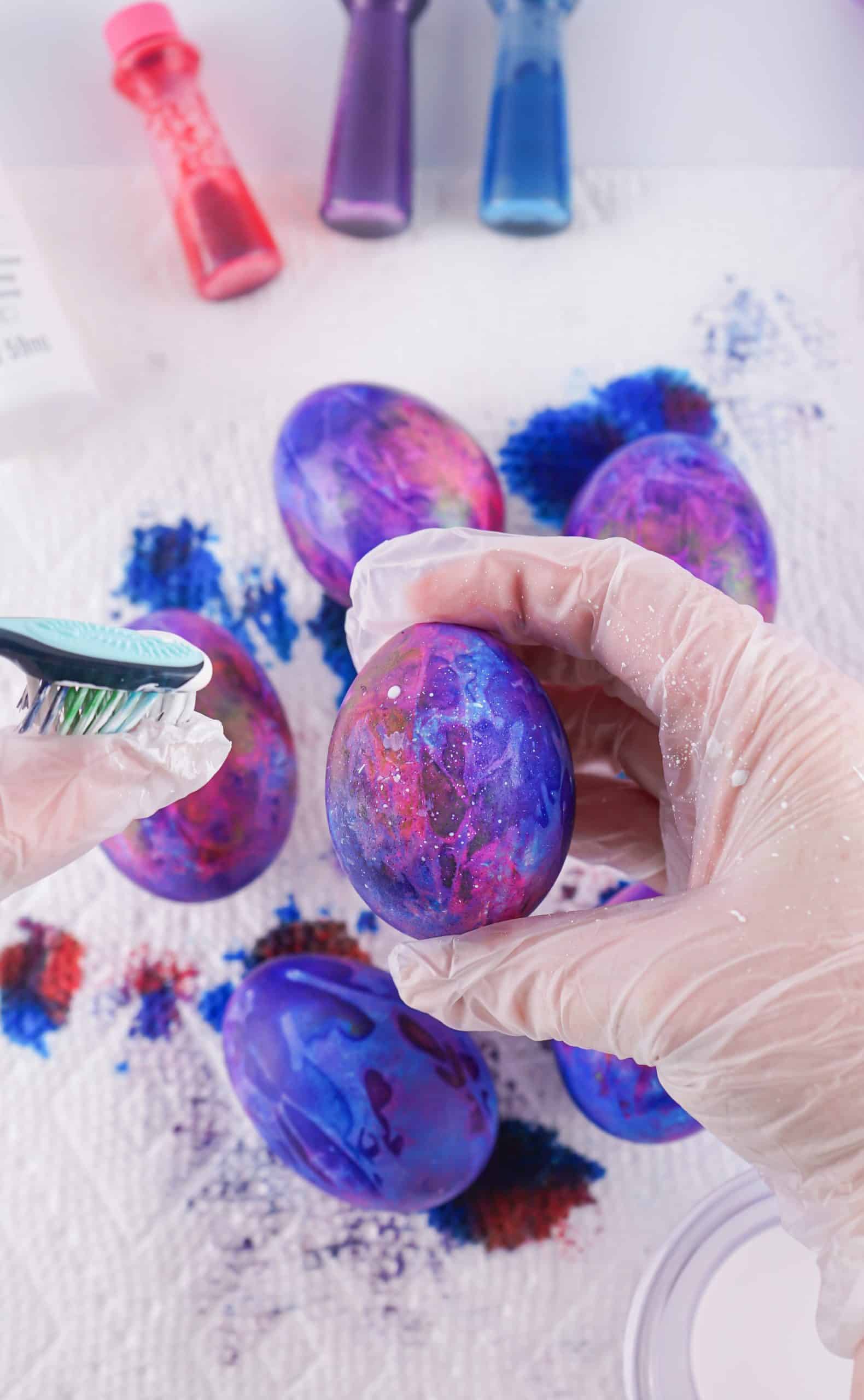a hand shown flicking white paint from a tooth brush onto dyed easter eggs