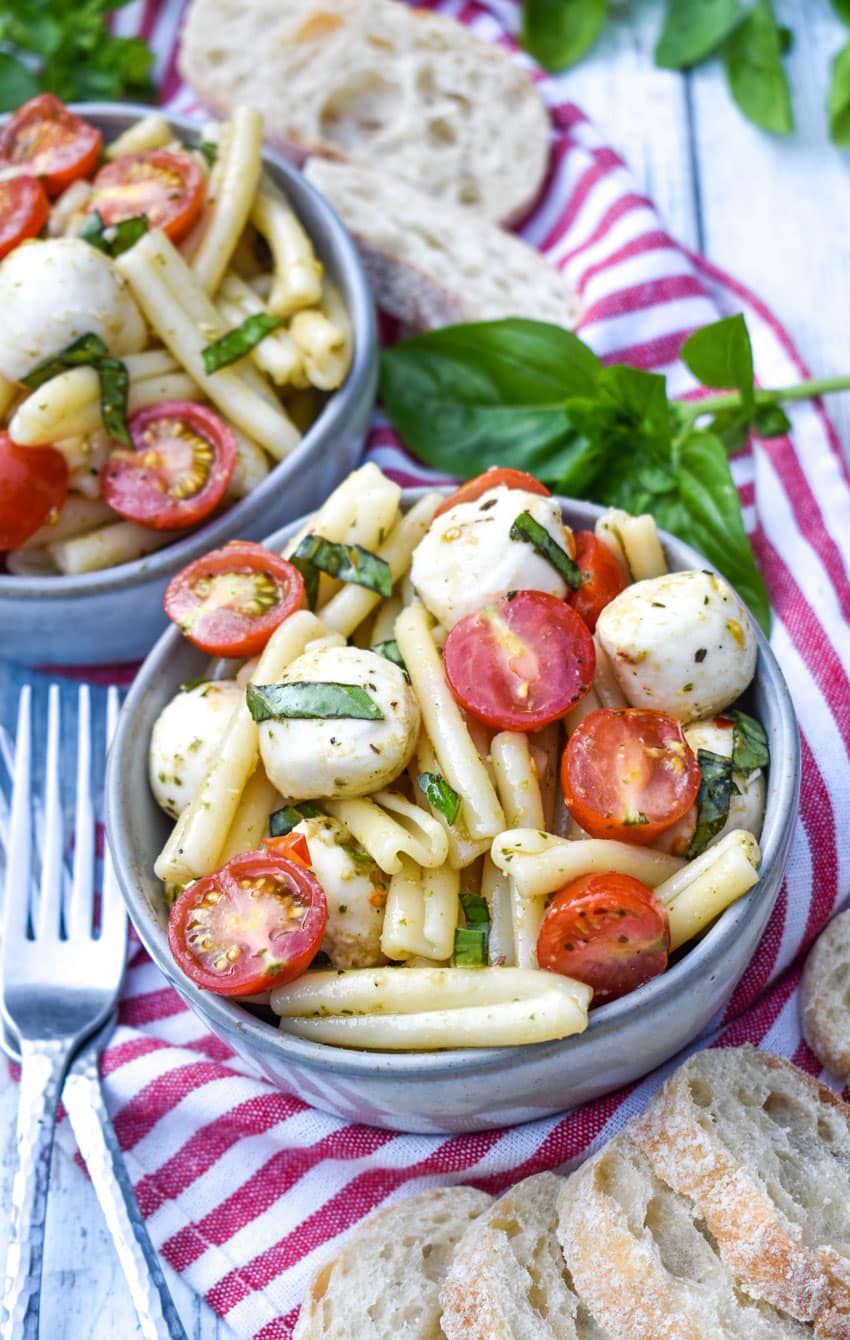 caprese pasta salad in two small gray bowls with fresh basil leaves and baguette slices on the side