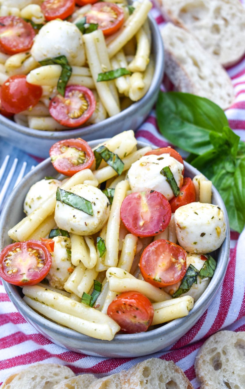 caprese pasta salad in two small gray bowls with fresh basil leaves and baguette slices on the side