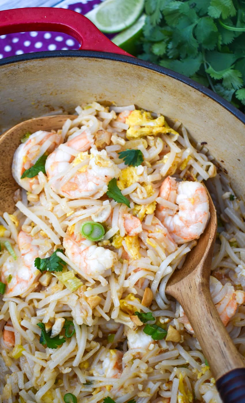 homemade shrimp pad thai being scooped out of a red dutch oven by wooden serving spoons