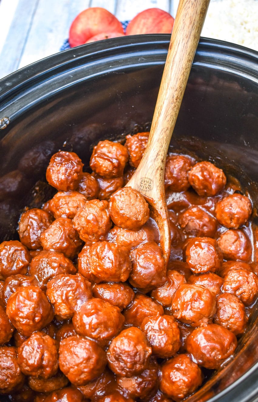 a wooden spoon scooping apple butter meatballs out of a black crockpot