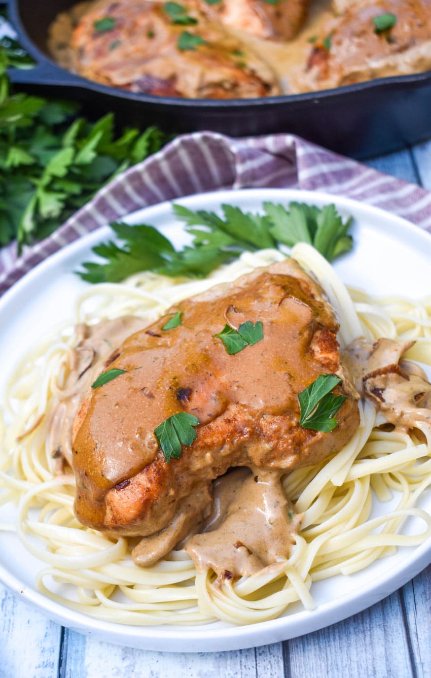 a piece of boursin chicken in cream sauce over cooked pasta on a white plate with fresh herbs on the side