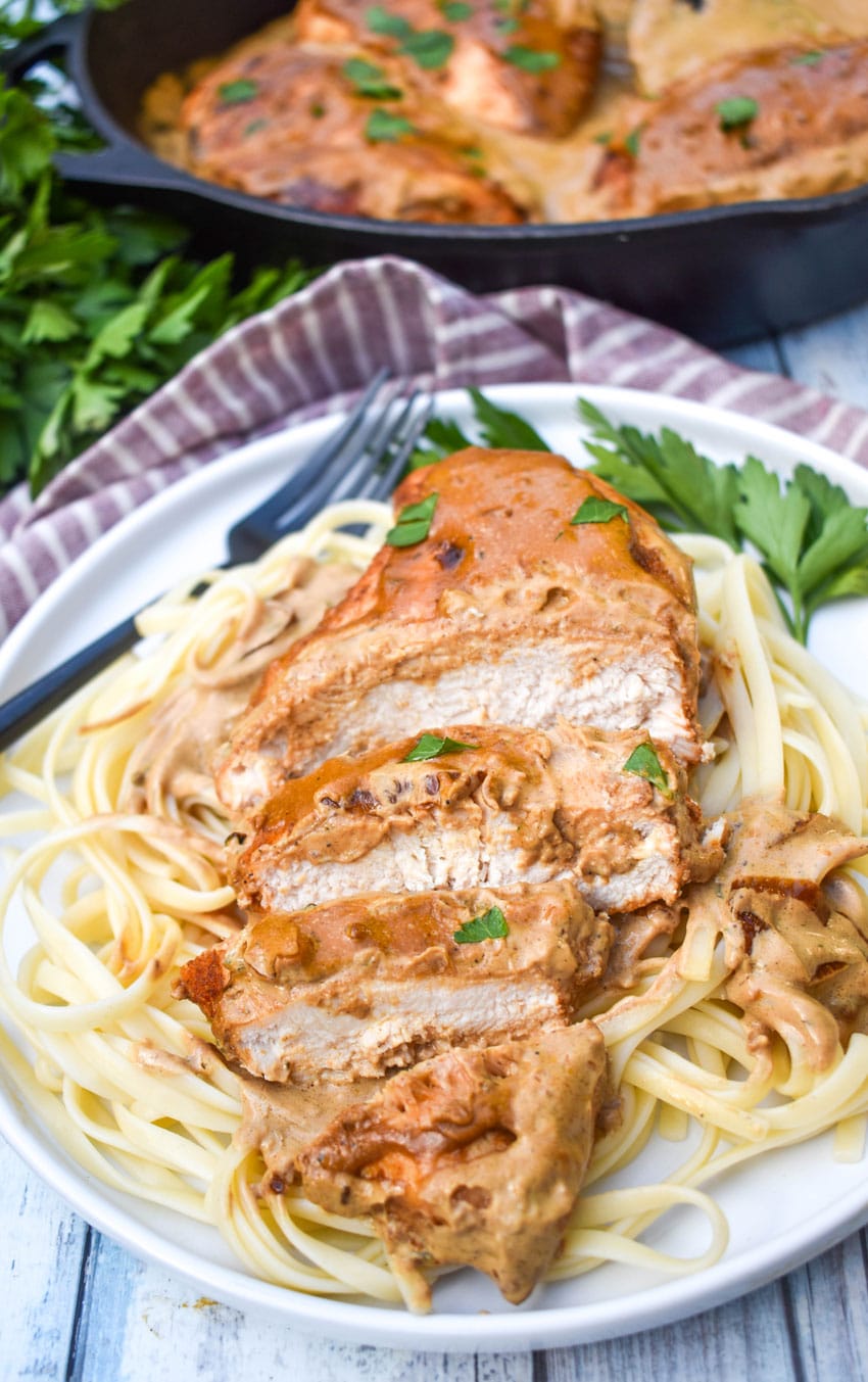 a sliced piece of boursin chicken in cream sauce over cooked pasta on a white plate with fresh herbs on the side