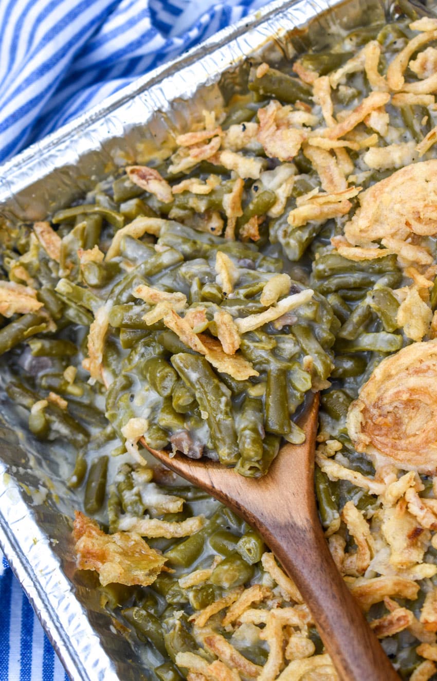 a wooden spoon scooping smoked green bean casserole out of a disposable aluminum pan