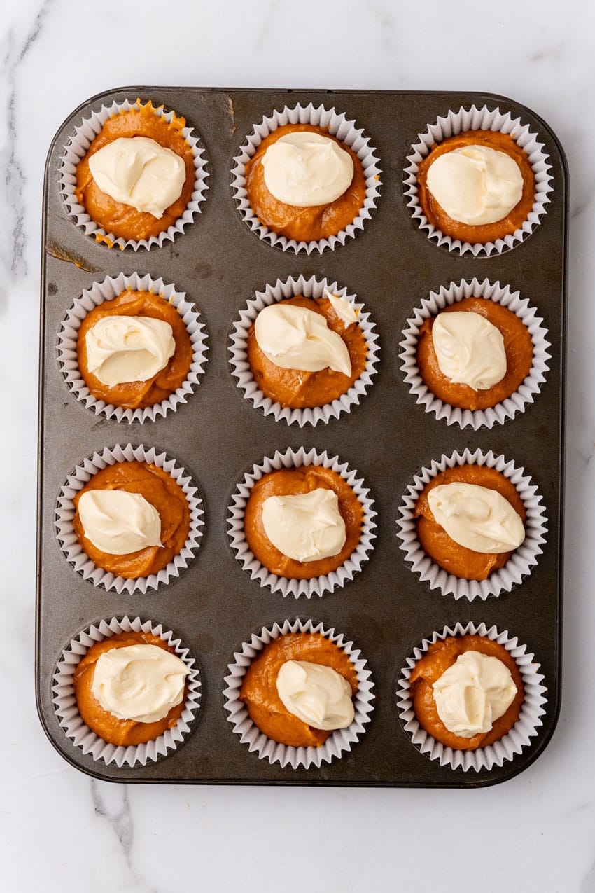 dollops of whipped cream cheese on top of pumpkin muffin batter in white cupcake liners in a metal muffin pan