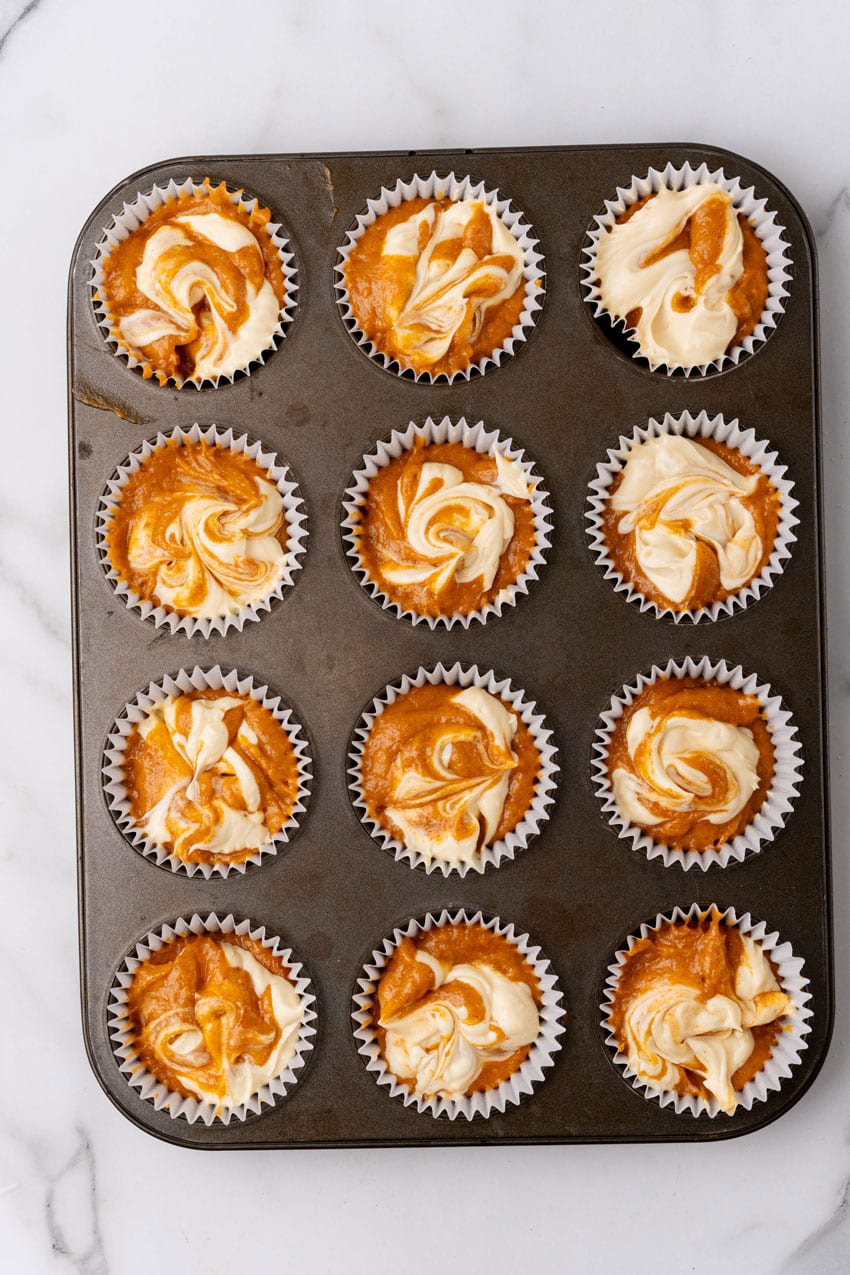 pumpkin cream cheese swirled muffin batter in white cupcake liners in a metal muffin pan