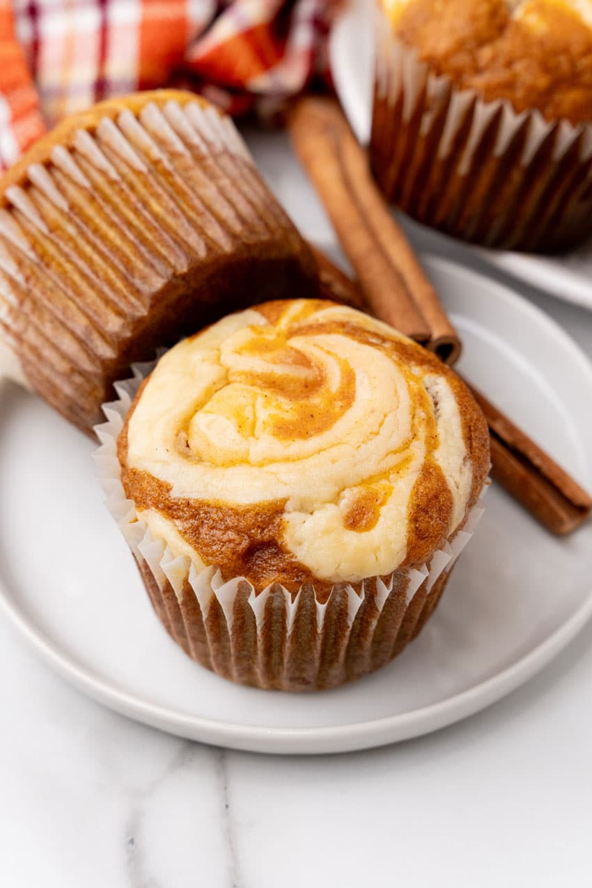 two pumpkin cream cheese swirl muffins on a small white plate with cinnamon sticks on the side