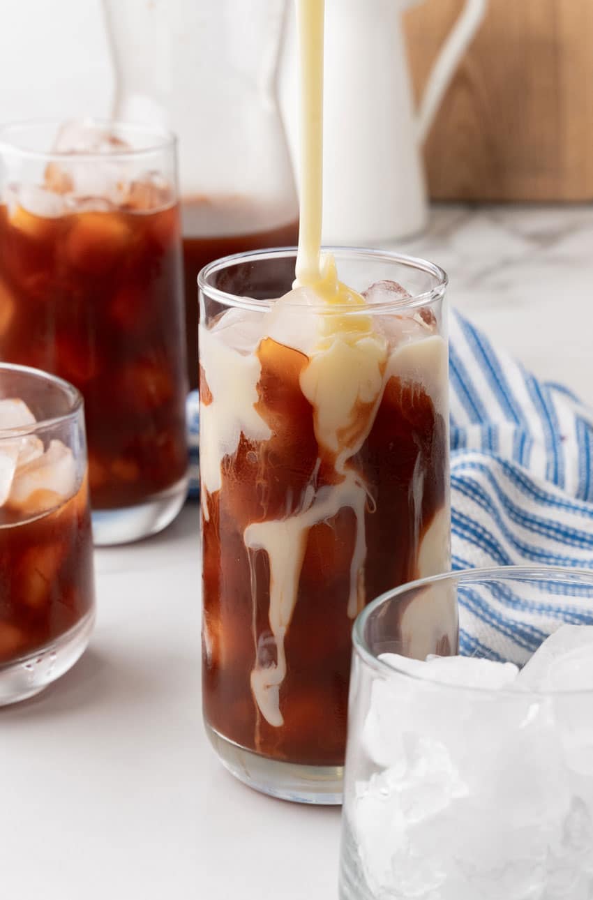 sweetened condensed milk being poured over ice into a glass filled with brewed tea