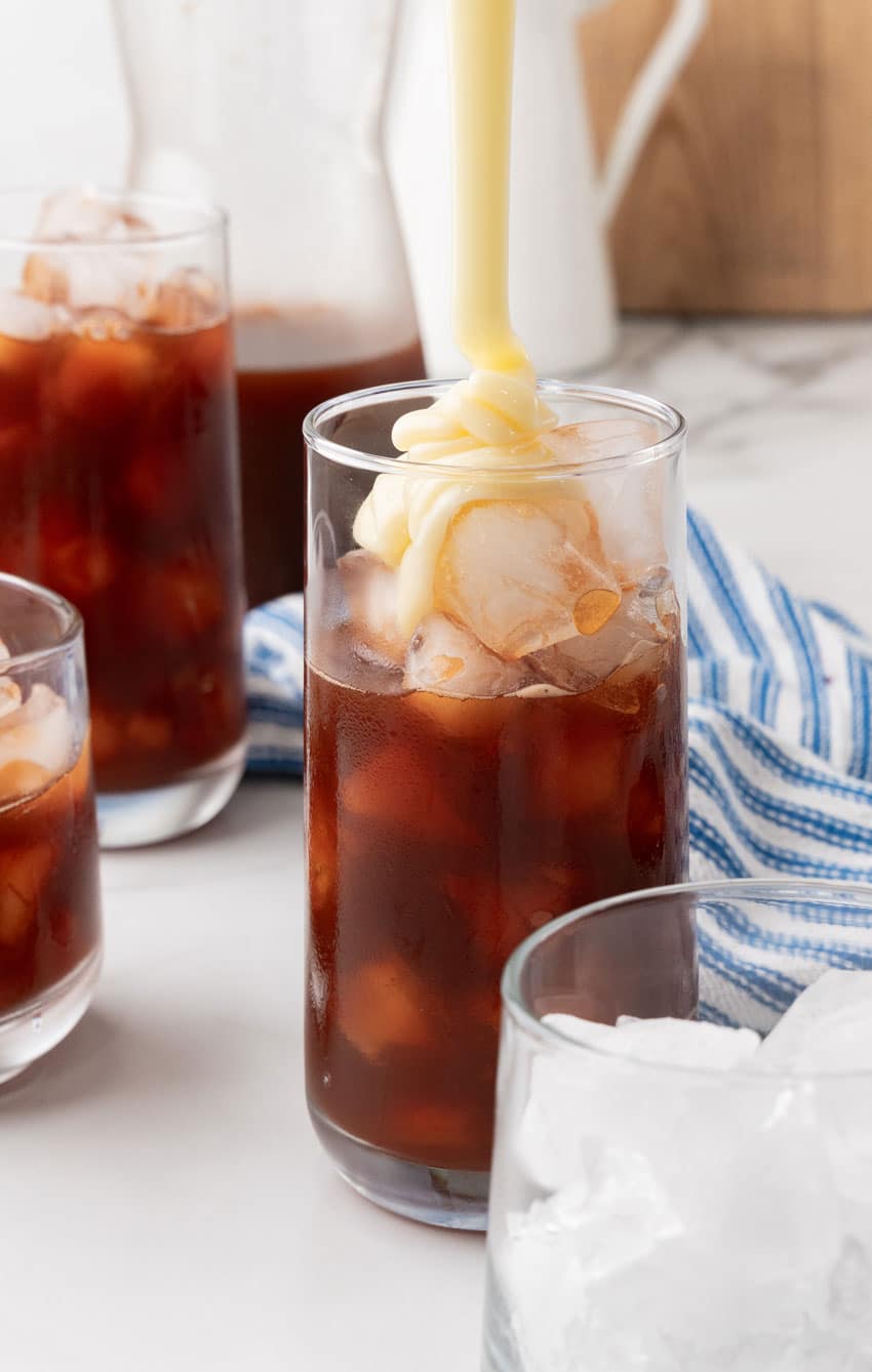 sweetened condensed milk being poured over ice into a glass filled with brewed tea