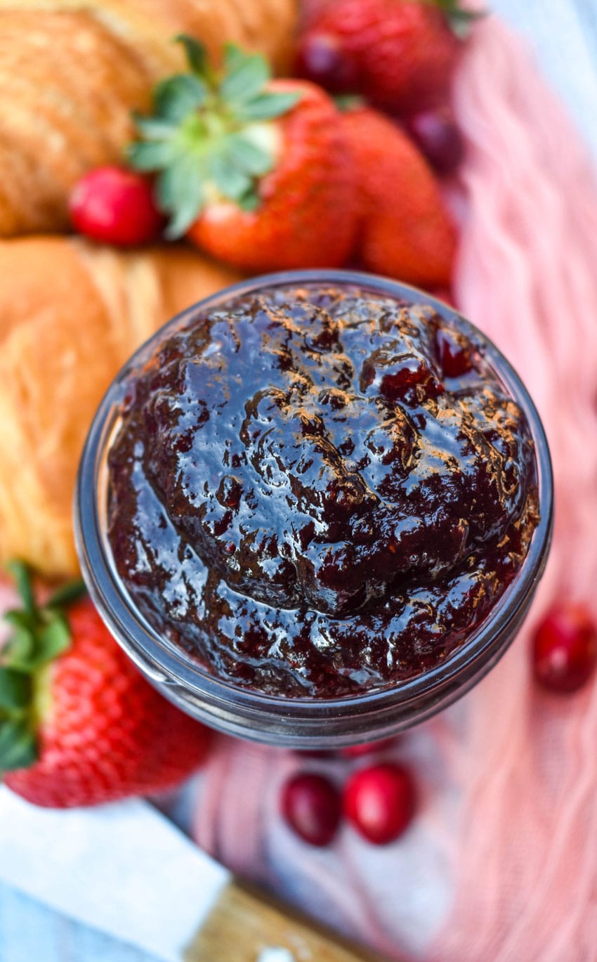 homemade christmas jam in a small glass jar surrounded by fresh berries and croissant rolls