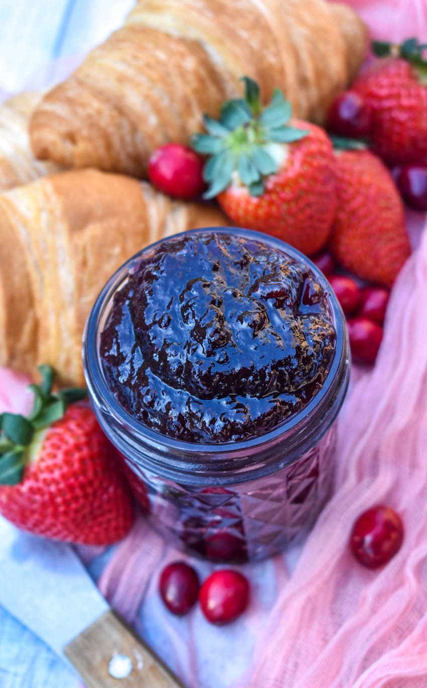 homemade christmas jam in a small glass jar surrounded by fresh berries and croissant rolls