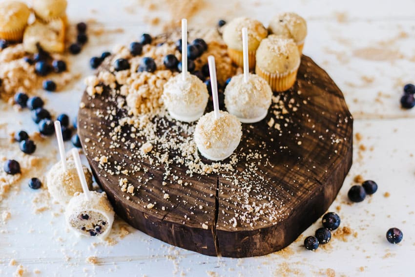 blueberry muffin cake pops on a wooden cutting board surrounded by graham cracker crumbs and fresh blueberries