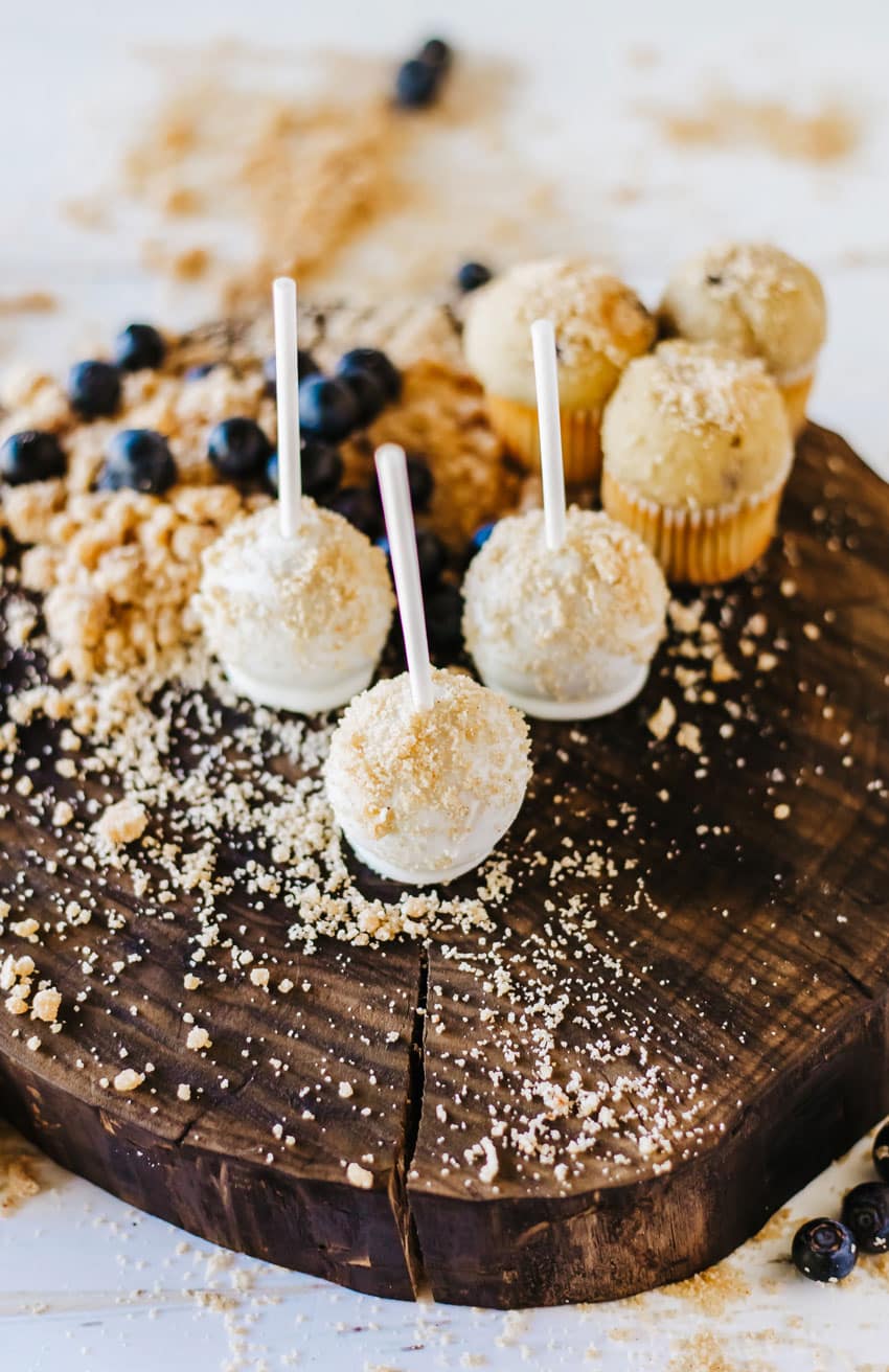 blueberry muffin cake pops on a wooden cutting board surrounded by graham cracker crumbs and fresh blueberries