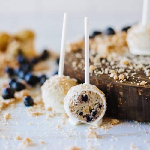 two halves of a blueberry muffin cake pop on a wooden table surrounded by graham cracker crumbs and fresh blueberries