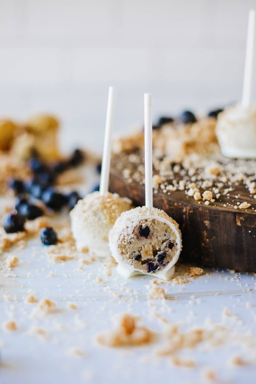 two halves of a blueberry muffin cake pop on a wooden table surrounded by graham cracker crumbs and fresh blueberries