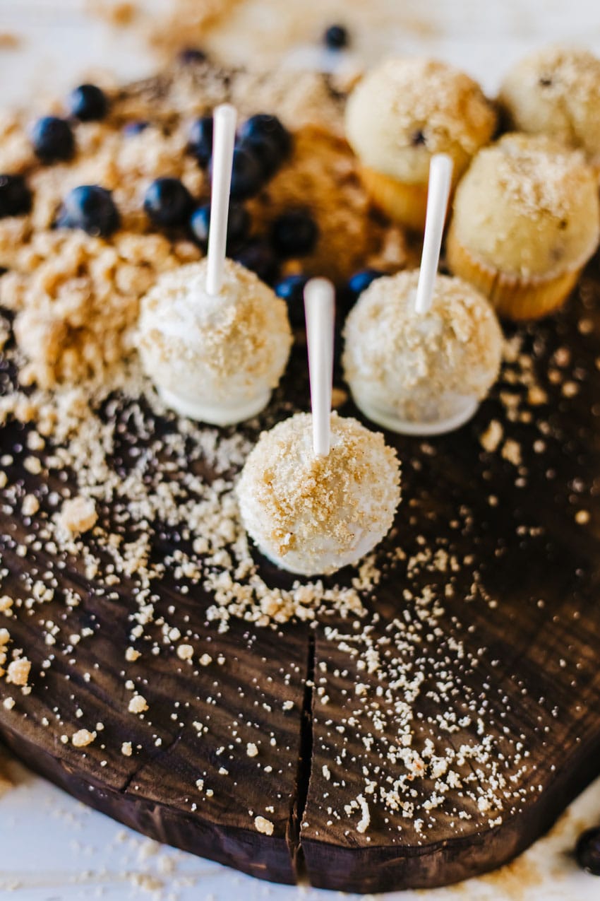 blueberry muffin cake pops on a wooden cutting board surrounded by graham cracker crumbs and fresh blueberries